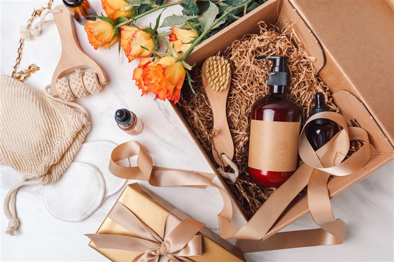 Uma caixa de presente aberta contendo itens de cuidados pessoais, como frascos de cosméticos com rótulos neutros, uma escova de madeira, um massageador manual, e flores laranjas. Ao redor, há um saco reutilizável de tecido, discos de algodão, fitas douradas e outro presente embrulhado em papel dourado com laço de fita. A cena sugere um kit de spa ou bem-estar, em um fundo branco de mármore.
