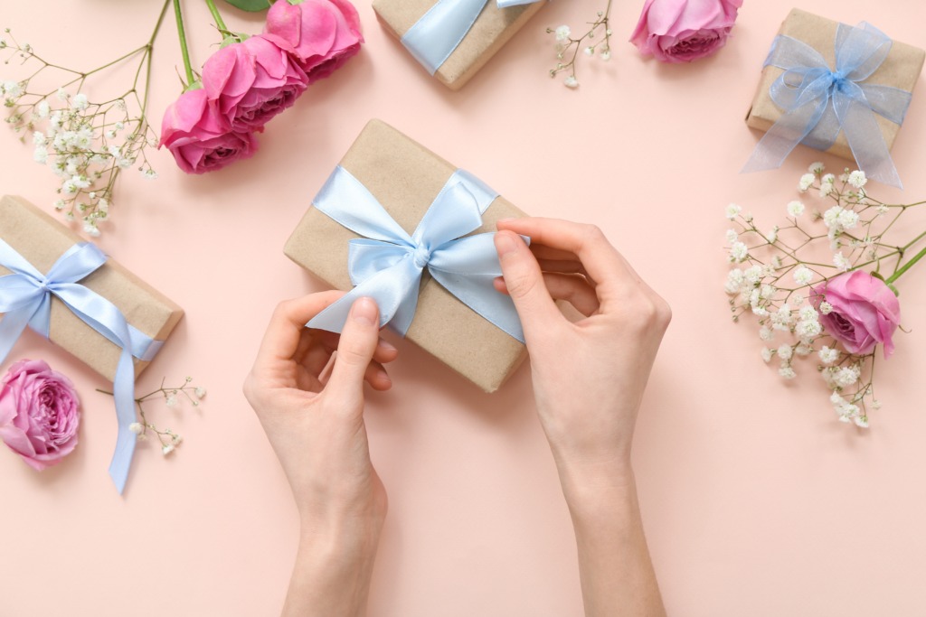 Duas mãos amarrando um laço azul-claro em um presente embrulhado com papel kraft, sobre uma superfície rosa-claro. Ao redor, há outros presentes semelhantes, rosas cor-de-rosa abertas, flores brancas delicadas e fitas de cetim azul.