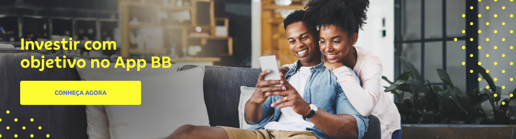 Um casal jovem, sorridente e de pele negra está sentado confortavelmente em um sofá moderno dentro de uma sala iluminada com plantas e móveis ao fundo. O homem segura um celular, enquanto a mulher, abraçada a ele por trás, observa a tela com interesse. Ele usa camisa jeans, blusa branca e bermuda marrom e ela casaco branco e calça jeans.
À esquerda da imagem, em letras amarelas, está o texto "Investir com objetivo no App BB". Abaixo, um botão: Conheça agora