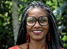 Fotografia de Aline Najara da Silva Congalves. Na imagem está com cabelo rastafari, óculos de grau com armação preta e brincos de sementes.