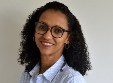 Rejane Mira. Mulher de camisa branca com detalhes em azul. Usa óculos e cabelos cacheados.