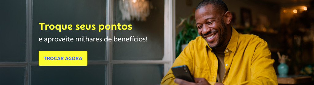 Homem negro, sorridente, usando camisa amarela, está sentado em um ambiente aconchegante e olha para o celular em suas mãos. Ao fundo, há uma janela e iluminação quente. À esquerda da imagem, o texto diz: Troque seus pontos e aproveite milhares de benefícios!'. Logo abaixo, há um botão amarelo com o texto em azul: TROCAR AGORA