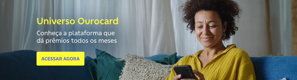 Mulher negra, de cabelo crespo curto, vestindo blusa amarela, está sentada em um sofá azul em um ambiente iluminado, olhando para o celular com expressão satisfeita. Ao fundo, há cortinas claras e almofadas. À esquerda da imagem, o texto diz: Universo Ourocard. Conheça a plataforma que dá prêmios todos os meses. Abaixo, há um botão amarelo com o texto em azul: ACESSAR AGORA